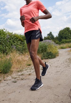 Person wearing coral shirt and black shorts running on a dirt path surrounded by green bushes and grass under a partly cloudy sky.