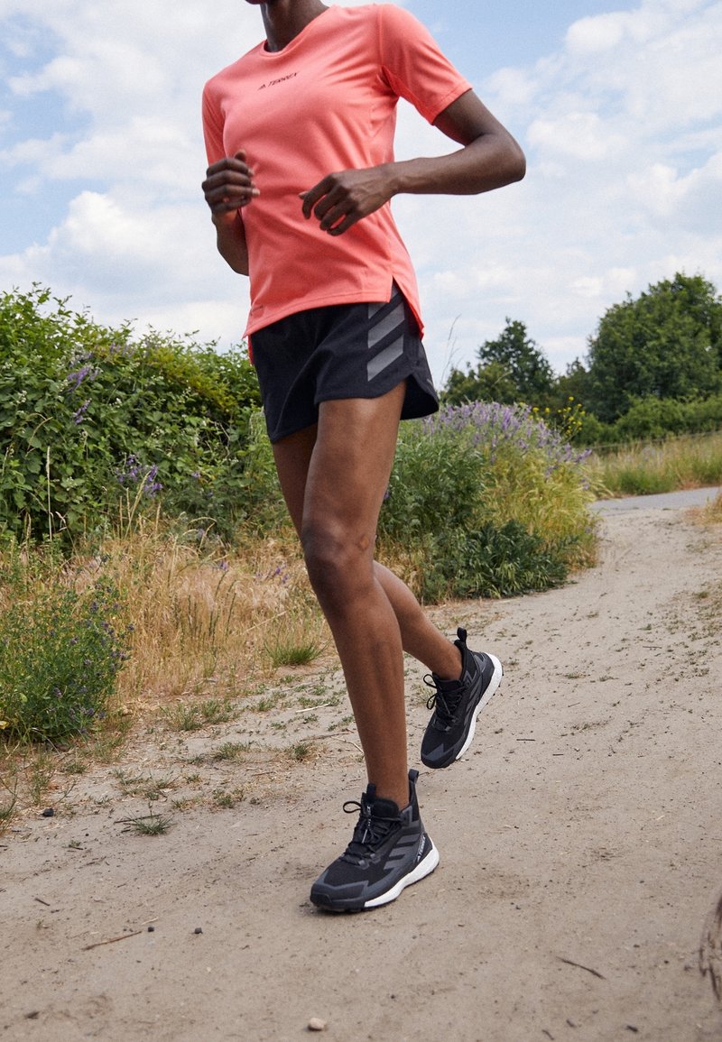 Personne portant un t-shirt corail et un short noir courant sur un chemin en terre entouré de buissons verts et d'herbe sous un ciel partiellement nuageux.