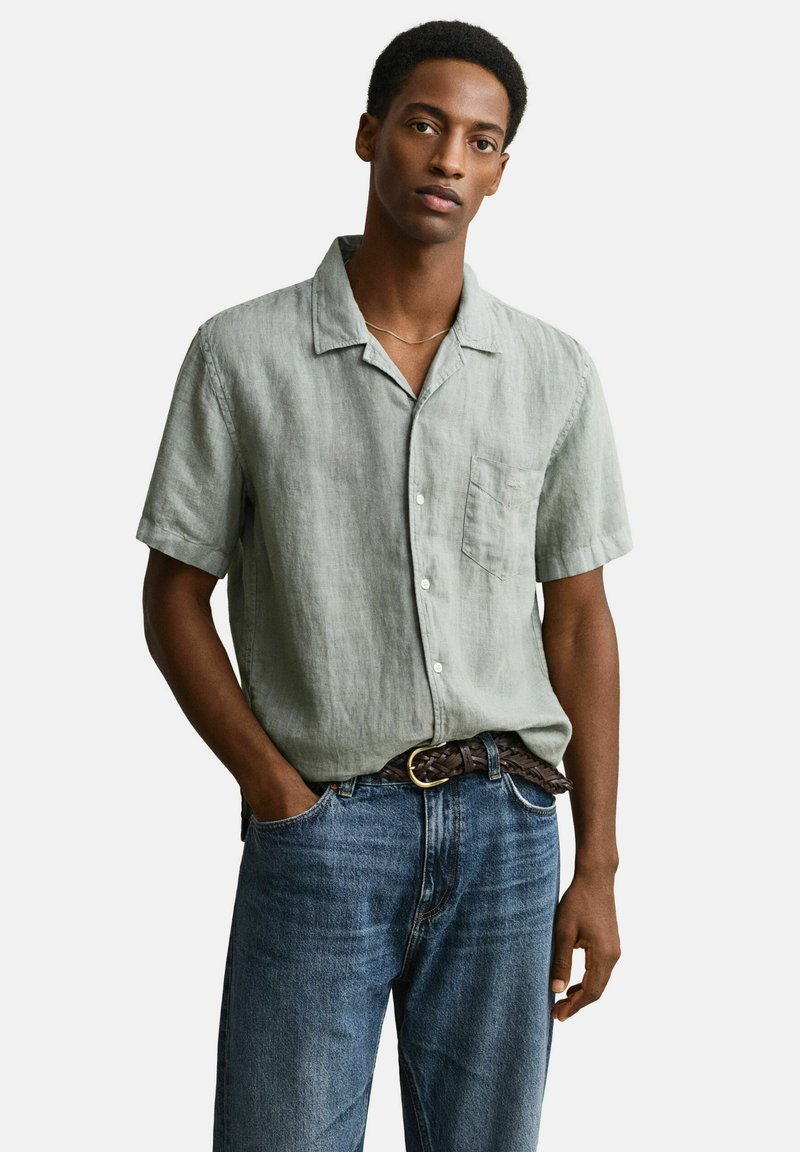 Uomo che indossa una camicia leggera a maniche corte grigio chiaro con bottoni, jeans blu e cintura intrecciata marrone, in piedi con una mano in tasca su sfondo bianco.