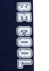 Tessuto blu navy con audace lettering bianco rovinato che scrive "BAGOOP" disposto verticalmente. Il design enfatizza un aspetto testurizzato e consumato.