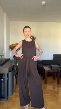 Γυναίκα με σκούρο μωβ ολόσωμη φόρμα χωρίς μανίκια στέκεται χαμογελαστή στο σαλόνι με πιάνο, φυτά, καναπέ και ξύλινο τραπέζι.