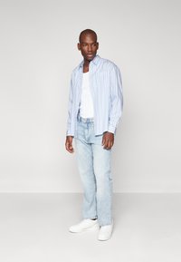 Camisa a rayas azul claro con cierre de botones, llevada sobre una camiseta blanca sin mangas. Combinada con jeans azul claro y zapatillas blancas. Textura suave.