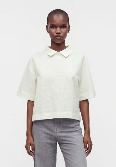 Personne debout de face, portant un haut blanc à manches courtes avec col et un jean gris, sur un fond uni et clair.