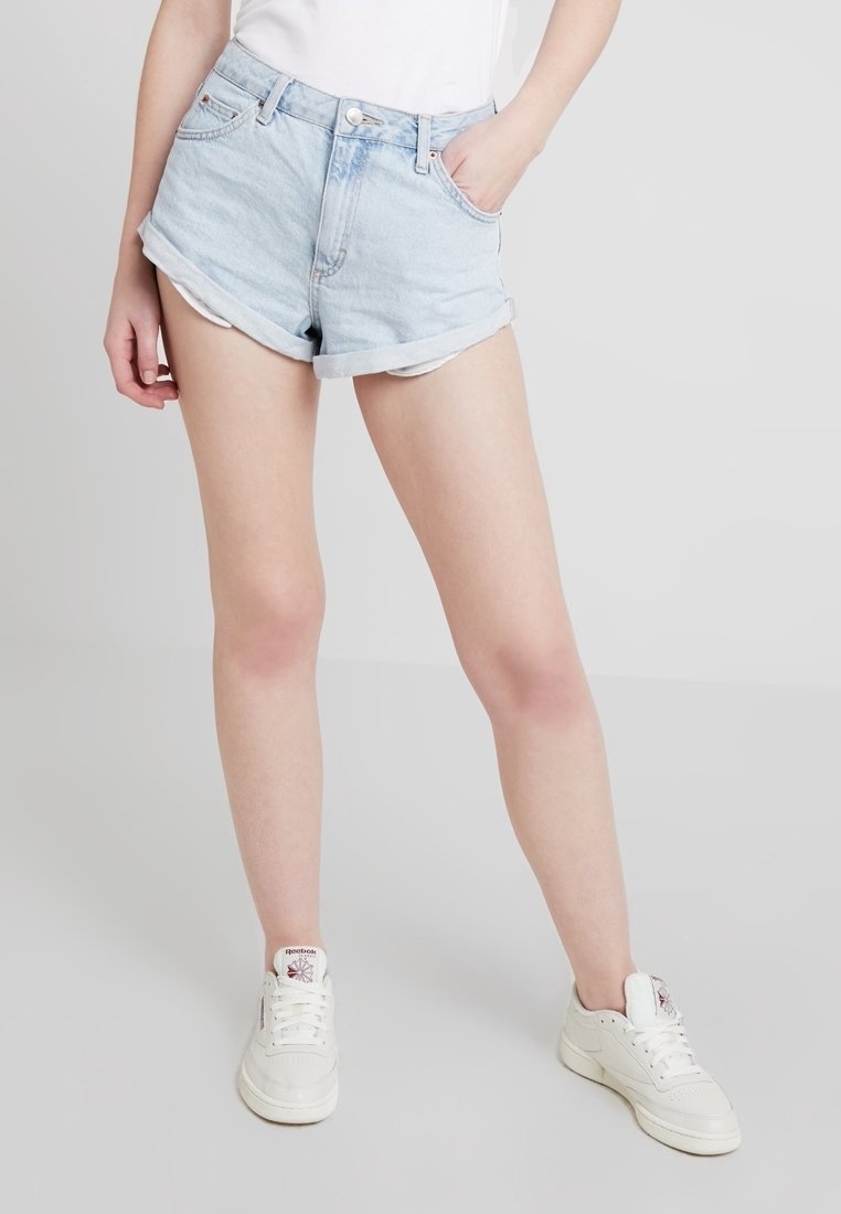 Persona que lleva pantalones cortos de mezclilla azul claro arremangados, zapatillas blancas y una blusa blanca, con una mano en el bolsillo, de pie sobre un suelo gris.