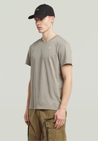 Jasnoszary bawełniany t-shirt z krótkimi rękawami i okrągłym dekoltem, zestawiony z khaki spodniami cargo z zamkami i pętlami, noszony z czarną czapką.