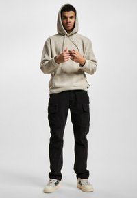 Beige snorrelue med snørebånd og frontlomme, kombinert med svarte cargo-bukser med sidelommer, iført hvite og grønne sneakers.