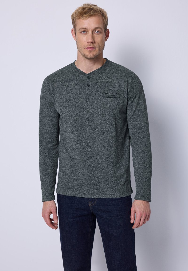 Graues Langarm-Henley-Shirt aus strukturiertem Stoff mit einer Drei-Knopf-Leiste und seitlichen Schlitzen, kombiniert mit dunklen Jeans.