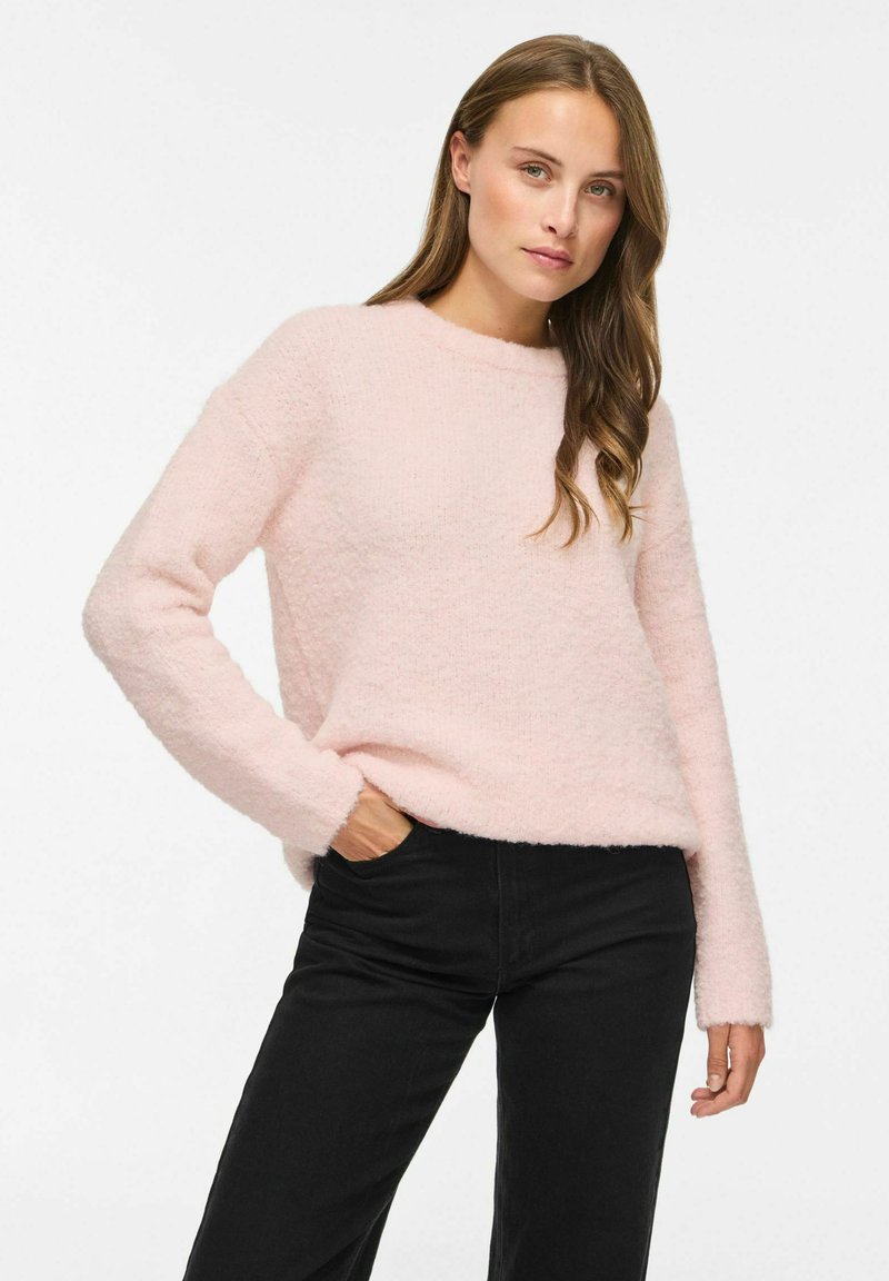 Femme aux longs cheveux portant un pull duveteux rose clair et un pantalon noir, posant avec une main dans la poche sur fond blanc.
