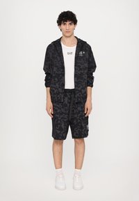 Svart treningstøy med en hettegenser med et subtilt mønster, matchende shorts og hvite sneakers med synlige sokker.