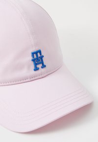Rosa cap laget av stoff med en blå logo sydd inn som har sammenflettede bokstaver. Designet inkluderer en buet skjerm og en strukturert krone.