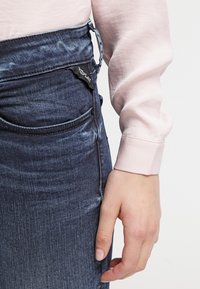 Vaqueros de mezclilla con un lavado azul oscuro y un ligero degradado. La cinturilla incluye un lazo decorativo, y se puede ver una camiseta de manga larga rosa claro.