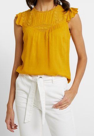 Mouwloze mosterdgele blouse met geborduurde inzet en gerimpelde schouders, gecombineerd met witte, hooggesneden broek met een strikband aan de voorkant.