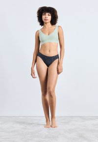 Világoszöld bralette széles pántokkal, sötét mintás bikini alsóval párosítva. A modell mezítláb áll egy texturált szürke felületen.