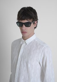 Camisa blanca de botones con patrones geométricos texturizados; combinada con gafas de sol negras angulares, sobre un fondo gris claro.
