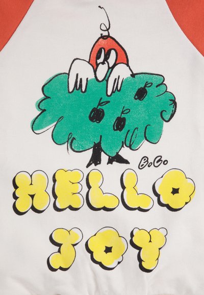Una tela blanca con mangas raglán rojas presenta un personaje de dibujos animados asomándose desde un arbusto verde. El texto debajo dice "HOLA ALEGRÍA" en letras gruesas amarillas.