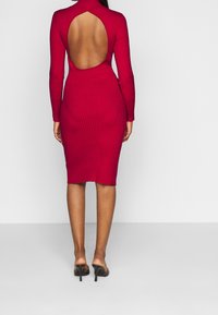 Ribbed red dress with long sleeves and a large back cut-out, fitted silhouette, knee-length, paired with black open-toe heels.