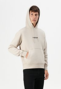 Hellbeige Hoodie mit einer Vordertasche, Kapuze mit Kordelzug und schwarzem Logo-Text auf der Brust. Hergestellt aus glattem Stoff mit gerippten Bündchen.