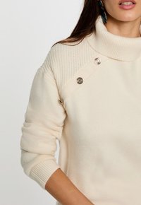 Cremfarbener Rollkragenpullover mit gerippter Struktur, glattem Körper, Strickdetails an den Schultern und metallischen Knopfakzenten.