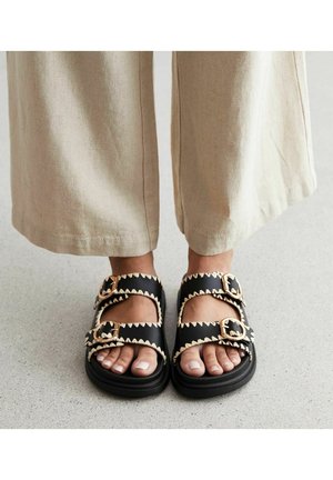 Czarne sandały z złotymi klamrami, zygzakowate wykończenie wzdłuż brzegów, płaska podeszwa, otwarty nos, noszone z beżowymi spodniami w szerokim kroju.