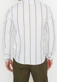 Valkoinen pitkähihainen paita, jossa pystysuorat harmaat raidat, napilliset hihansuut ja pyöristetty helma. Valmistettu teksturoidusta kankaasta, esittelee rentoa muotoilua.