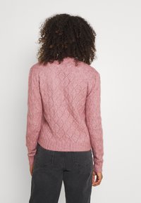 Osoba s kudrnatými vlasy má na sobě růžový svetr s diamantovým vzorem a tmavě šedé džíny, zobrazená zezadu na bílém pozadí.