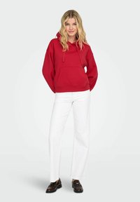 Jeune femme debout portant un sweat à capuche rouge, un pantalon blanc et des mocassins marron foncé, devant un fond gris clair uni.