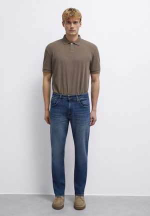 Junger Mann mit kurzen lockigen blonden Haaren trägt ein braunes Poloshirt, blaue Jeans und beige Wildlederschuhe, steht vor einer weißen Wand.
