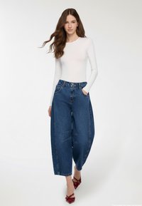 Top bianco a maniche lunghe aderente abbinato a jeans blu a gamba larga con vita alta e cuciture in vista, completato da tacchi rossi.