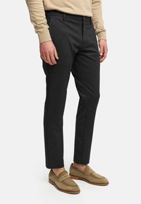 Pantalones negros a medida hechos de una tela suave, con un ajuste slim, bolsillos frontales y cremallera. Combinados con mocasines de ante color marrón.
