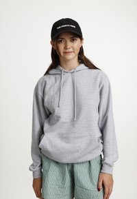 Grijze hoodie gemaakt van zachte stof, met een kangoeroezak en trekkoorden. Gecombineerd met groene gestreepte shorts en een zwarte pet.