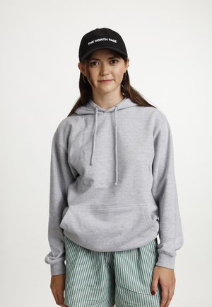 Grauer Kapuzenpullover aus weichem Stoff, mit einer Kängurutasche und Kordeln. Kombiniert mit grün gestreiften Shorts und einer schwarzen Kappe.