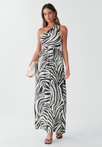 Sort-hvid zebra print maxikjole med én skulder, med en rynket talje og flydende skørt. Model holder en flettet clutch taske og har sorte sandaler på.