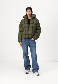 Olivenfarget dunjakke med en boksaktig design, høy krage og kviltet mønster. Kombinert med løstsittende blå jeans og lyse sneakers.