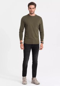 Vihreä oliivinvihreä pitkähihaisessa paidassa, slim-fit mustissa farkuissa ja vaaleissa harmaissa lenkkareissa. Sileä kangas ja yksinkertainen muotoilu, ei näkyviä kuvioita.