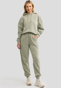 Helle grüne Kapuzenjacke mit Kordeln und passender Jogginghose. Weicher Stoff, lockere Passform und elastische Bündchen. Tragen mit beigen Sneakers.