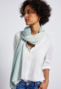 Écharpe bleu clair avec un motif en grille texturé, drapée autour du cou. Fabriquée en tissu doux, associée à une chemise blanche à boutons et un jean en denim.