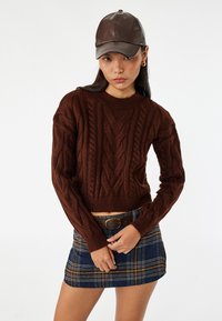 Pull en mailles marron à manches longues et col rond, associé à une jupe mini à carreaux bleus et une casquette marron.