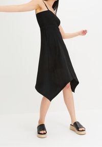 Černé bezrukávové šaty s texturovaným živůtkem, asymetrickým lemem a volným střihem. Doplněné černými skluznými sandály.