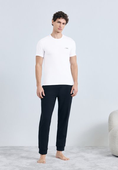 Hvid T-shirt med korte ærmer og et lille logo på brystet, kombineret med sorte joggebukser. Begge dele er lavet af blødt, glat stof.