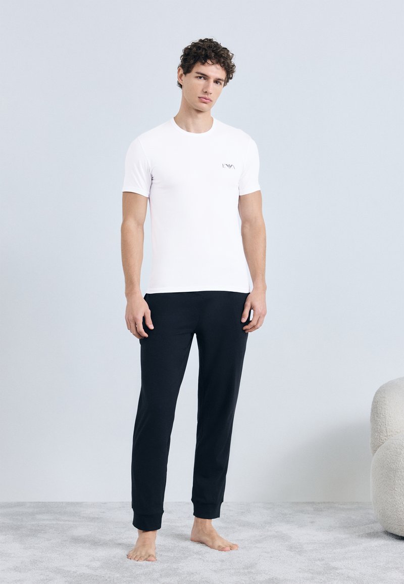 Balta īsroku t-krekls ar mazu logotipu uz krūtīm, kopā ar melnām sporta biksēm. Abas drēbes izgatavotas no mīksta, gluda auduma.