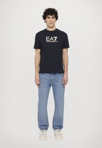 Temno modra majica s kratkimi rokavi z belim logotipom "EA7 Emporio Armani", v kombinaciji s svetlo modrimi džinsom in kremnimi supergami.