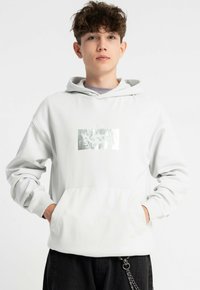 Biała bluza z kapturem wykonana z miękkiego materiału, z kieszenią z przodu oraz metalicznym nadrukiem na klatce piersiowej. Długie rękawy z ściągaczami.