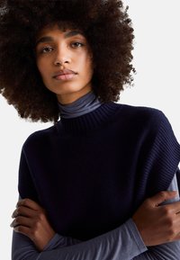 Mladá žena s přirozeně kudrnatými vlasy, oblečená v tmavě modrém svetru bez rukávů přes šedý rolák, s překříženýma rukama a neutrálním výrazem.