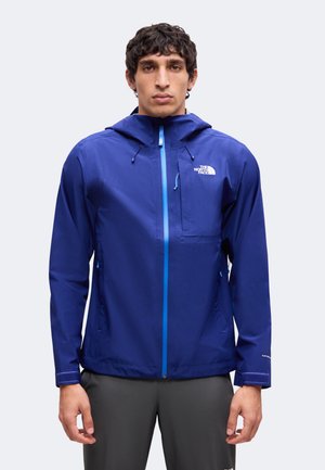 Homme portant une veste imperméable bleue The North Face avec capuche et poches zippées, debout devant un fond clair uni.