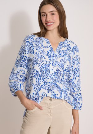 Nuori nainen hymyilee, pukeutunut valkoiseen paitapuseroon, jossa on sinisiä paisley-kuvioita, ja beigen värisiin housuihin, seisoo käsi taskussa.