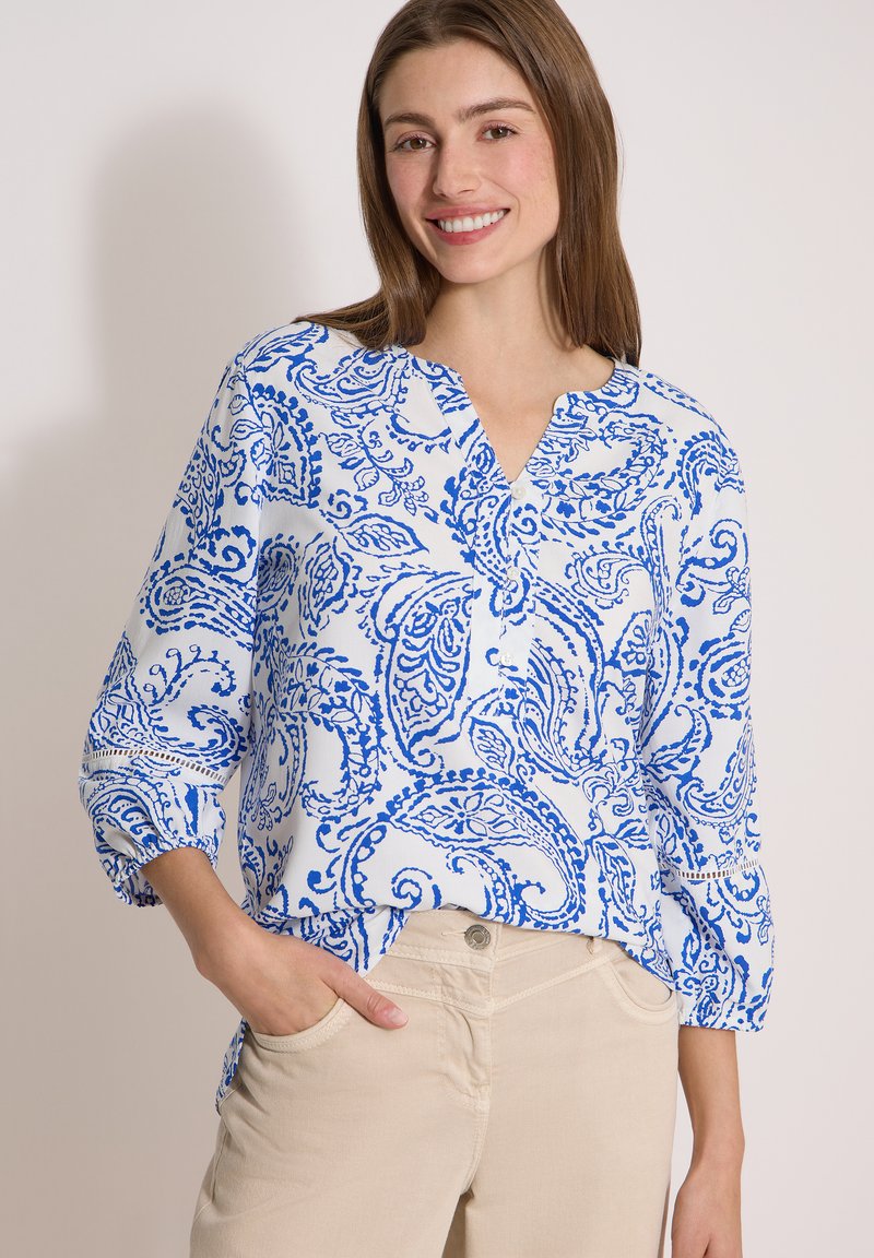 Jeune femme souriante, portant un chemisier blanc avec des motifs paisley bleus et un pantalon beige, debout avec une main dans la poche.