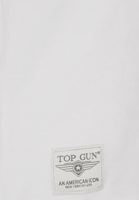 Hvid bomulds t-shirt med en grå label med "TOP GUN" og et vingen emblem. Glat tekstur, rund halsudskæring og korte ærmer.
