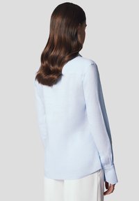 Jasnoniebieska koszula z długim rękawem, wykonana z miękkiego materiału, z kołnierzykiem zapinanym na guziki, zaokrąglonym dołem i mankietami zapinanymi na jeden guzik. Zobaczona z tyłu.