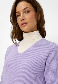 V-Ausschnitt Pullover in Lavendel mit geripptem Design. Über einem cremefarbenen Rollkragenpullover getragen, mit lockerer Passform und weichem Strickmaterial.