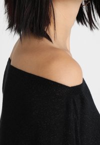 Svart off-shoulder-tröja med lös passform, med en bred halsringning och texturerad stickning. En subtil glans från garnet är synlig.